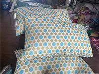 Photo showing three stacked feather filled throw pillows with a geometric pattern in blue, green and beige on a white background resting on a wooden floor.