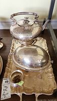 Photo showing the tea pot with decorative engraved patterns, the serving dish with a domed lid, the waste bowl, all placed on a large silver tray with ornate engraved design and scalloped edges.