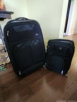 Front view showing both Skyway and Fai black suitcases side by side on wooden floor.