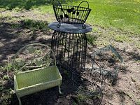 Photo of all items showing the black metal table with round top and four metal baskets/shelves around it on grass outdoors.