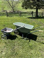 Side view of the green Turfx metal wheelbarrow and gray Promix fertilizer spreader on grass with wooden fence and trees in background.