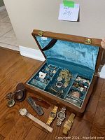 Open wooden jewelry box displaying blue velvet interior with multiple jewelry pieces and watches inside as well as some items placed around the box on wooden floor.
