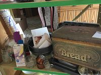 Photo showing vintage metal bread box labeled 'Bread' filled with various cookie cutters, utensils, and glass ramekins on a green shelf.