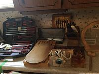 Photo showing multiple wooden cutting boards, a cheese tray, vintage carving sets in original cases, a wooden banana hanger, and a wooden block with cutters on a kitchen counter
