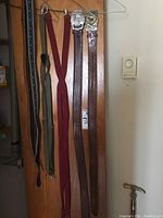 View of two leather belts with embossed designs and decorative metal buckles, alongside two pairs of suspenders (burgundy and olive green) hanging on a wooden door.