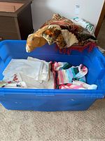 Blue plastic tote filled with fabric items including white linens, floral wall hanging, and multi-colored crochet cloth