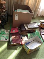 Wide view of various books loosely organized in boxes and piles on carpet near window with good natural light.