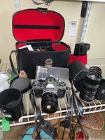 Photo of Canon AE-1 camera, lenses, flash, user manual, and carrying case displayed on wire shelving. Shows main camera body with attached 55mm lens, multiple lenses, flash unit in red and black case, and AE-1 manual.