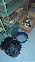 Two large enamel canning pots, one black with wire basket insert, and assorted glass jars in cardboard boxes on a basement floor under a metal shelving unit.