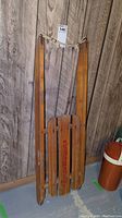 Wooden child's alpine glider sled leaning against a wood paneled wall, showing rope and construction details