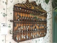 Full view of wooden rack with 42 assorted collector spoons hanging in two rows, carved mountain cabin scene on top.
