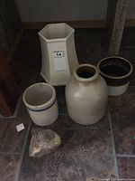 Overview of the lot showing five items: one large ceramic crock, two smaller crocks with blue rims, hexagonal cream-colored vase, and a rough rock on tiled floor.