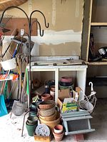 Photo showing collection of terracotta and plastic pots, wood bushel baskets stacked, black iron shepherd hooks, white metal hanging plant hanger, garden sprayer boxed, and garden hand tools against a garage wall.