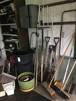 Photo showing several long-handled garden tools including shovels, rake, cultivator, and garden fork against a wall with a black plastic waste bucket and different planters nearby