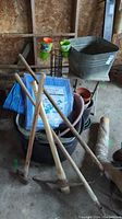 Photo showing wooden handled gardening tools including pickaxe, hoe, sledge hammer, blue tarp, metal wash tubs, and plastic planters stacked inside tubs.