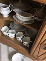 Photo showing multiple pieces of Johnson Bros dish set including cups, saucers, gravy boat, covered serving dish and plates stacked inside a wooden cabinet.