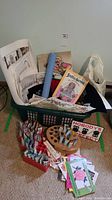 Photo showing a laundry basket filled with various liquid embroidery supplies including tubes of Tri-Chem liquid embroidery paints in a wooden rack, multiple pattern books including a stuffed animal pattern, and fabric remnants.