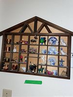 Full view of wooden display shelf with triangular roof shape mounted on wall, showing clear arrangement of miniature items including blue and white ceramic dishes and animal figurines.