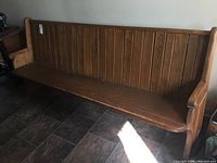 Full frontal view of a long wooden church pew on a tiled floor, showing the slatted backrest, seating surface, and armrests.