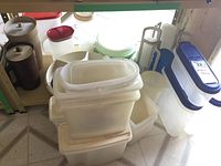 Photo of vintage plastic containers including translucent rectangular boxes with white hinged lids, blue and white containers, brown cylindrical containers, and a white measuring container on tiled floor.