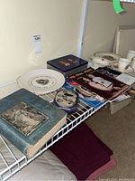 Photo showing assortment of plates, magazines, a blue tin, historic book, and bowls on a wire shelf