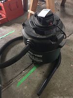 Full view of the Shop-Vac unit showing the cylindrical vacuum body in black/dark green, the attached hose, power cord wrapped around the top, and caster wheels.