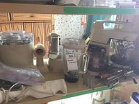 Photo showing cream-colored Oster stand mixer with multiple attachments and accessories including beaters and metal bowl, vintage food processor, and blender jar on two-tier shelf.