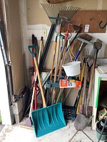 Photo shows collection of yard tools in garage corner including shovels, rakes, pitchfork, broom, hoe, hand tiller, hanging white planter, sponge mops, green plastic snow shovel, and parts of a tool rack.