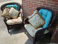 Two brown wicker armchairs with blue and patterned cushions against a brick wall, outdoors on concrete floor.