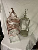 Three metal bird cages in rose, brownish-green, and white colors arranged on a white fabric background.