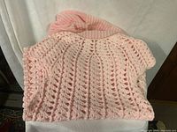 Photo showing folded light pink knitted baby dress with open stitch design and matching hooded cape placed on a white fabric surface.