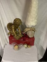 Full lot showing cherub figurine on red gift box, white faux Christmas tree with burlap base, Santa face plastic ornament, and three glass ornaments arranged below.