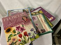 Photo of seven craft books stacked and fanned out showing colorful floral covers and titles emphasizing flower painting.