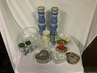 Photo showing assorted home decor items on a white cloth with blue candle holders, glass fishbowl, ceramic planters and vases, decorative metal trinket boxes in heart shapes, and other small decorative objects.