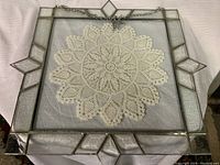 Full frontal image of square stained glass panel displaying crochet doily fixed in the center, metal chain attached at top for hanging.
