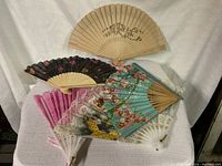 Five decorative hand fans arranged on a white fabric background showing various colors and designs.