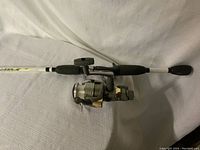 Fishing rod and reel laid flat on white fabric surface showing the entire rod and attached reel.