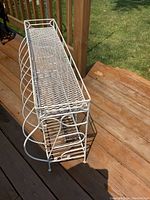 White metal flower stand shown outdoors on wooden deck from side angle to display lattice top, scrollwork sides, and overall shape