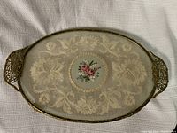 Photo of an oval glass dresser tray with ornate metal handles, gold leaf decorative pattern, and central floral motif. Visible crack noted on the glass.