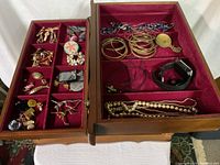View of open wooden jewelry box showing red velvet lined compartments filled with assorted jewelry items including bracelets, necklaces, cuff links, earrings, pins, and a watch.