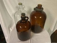 Three glass jugs displayed: two amber-colored and one clear. The largest amber jug with metal handle and clamp closure is at the back right. The smaller amber jug with cork stopper and loop handle is in front. The clear jug with metal screw-on lid and neck loop handle is at back left.