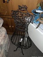 Photo of black decorative metal dress form with ornate wire design and flared base standing on carpeted floor beside furniture.