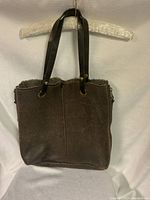 Front view of the brown UGG tote bag with leather handles hanging on a padded hanger. Shows the soft textured brown material and slightly furry top trim.