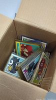 Top-down view of an open cardboard box filled with sleeved football cards showing multiple team colors and card backs.