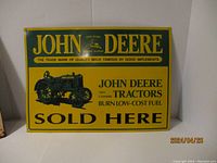 Photo of the embossed metal sign showing John Deere branding, tractor image, and promotional text with dimensions approximately 15 x 11.5 inches.