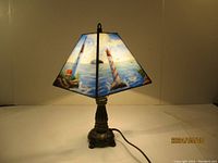 Front view of lamp showing lighthouse stained glass shade illuminated, metal base, power cord visible on surface.