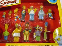 Close-up image of the 12 Simpsons figurines in molded plastic packaging, clearly showing details and characters.