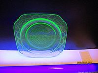 Photo of 7.5 inch square uranium depression glass plate glowing bright green under UV light, showing embossed decorative pattern.