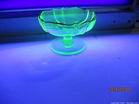 Photo showing the bright green fluorescence of the uranium glass bowl under UV light, highlighting the shape, scalloped edges, and pedestal base.