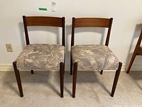 Front-facing photo of two teak dining chairs with fabric cushioned seats showing the front seat and legs.